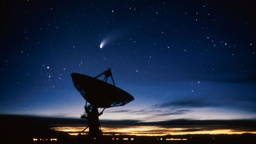 a bright comet in the sky over a large antenna dish