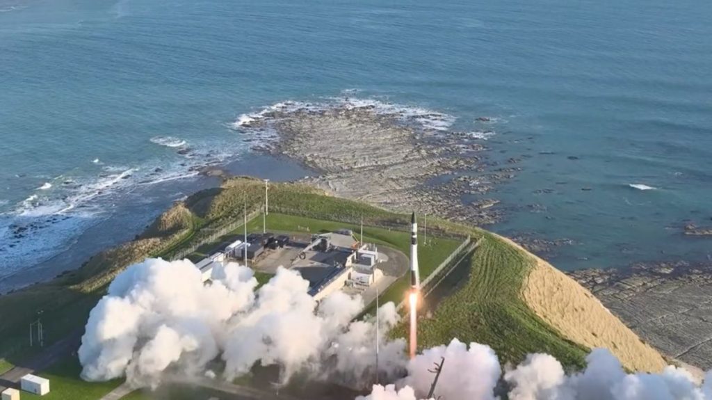 a white and black electron rocket lifts off from a seaside pad in new zealand with the ocean visible in the background.