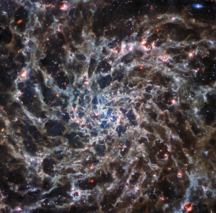 Gambar galaksi spiral dari James Webb Space Telescope.  Spiral terlihat seperti jaring laba-laba dengan daerah gas berwarna merah muda tersebar di seluruh gambar.
