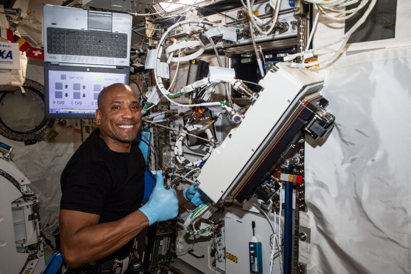 Seorang astronot berdiri di depan rak peralatan yang rumit.