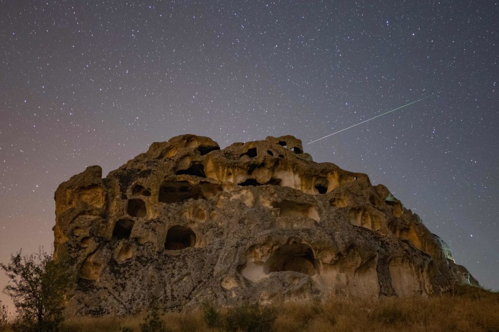 Kereta meteor Perseid melintasi langit di atas struktur batu besar dengan banyak gua dan lubang yang terukir.
