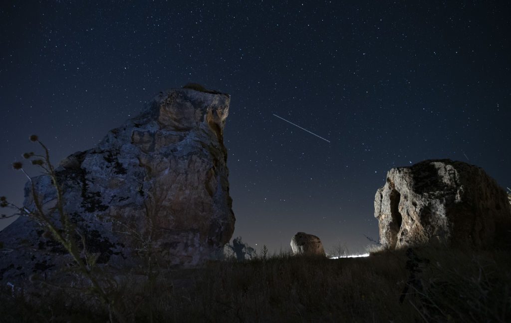 Kereta meteor yang panjang membentang melintasi langit di antara dua bangunan batu besar di latar depan gambar.