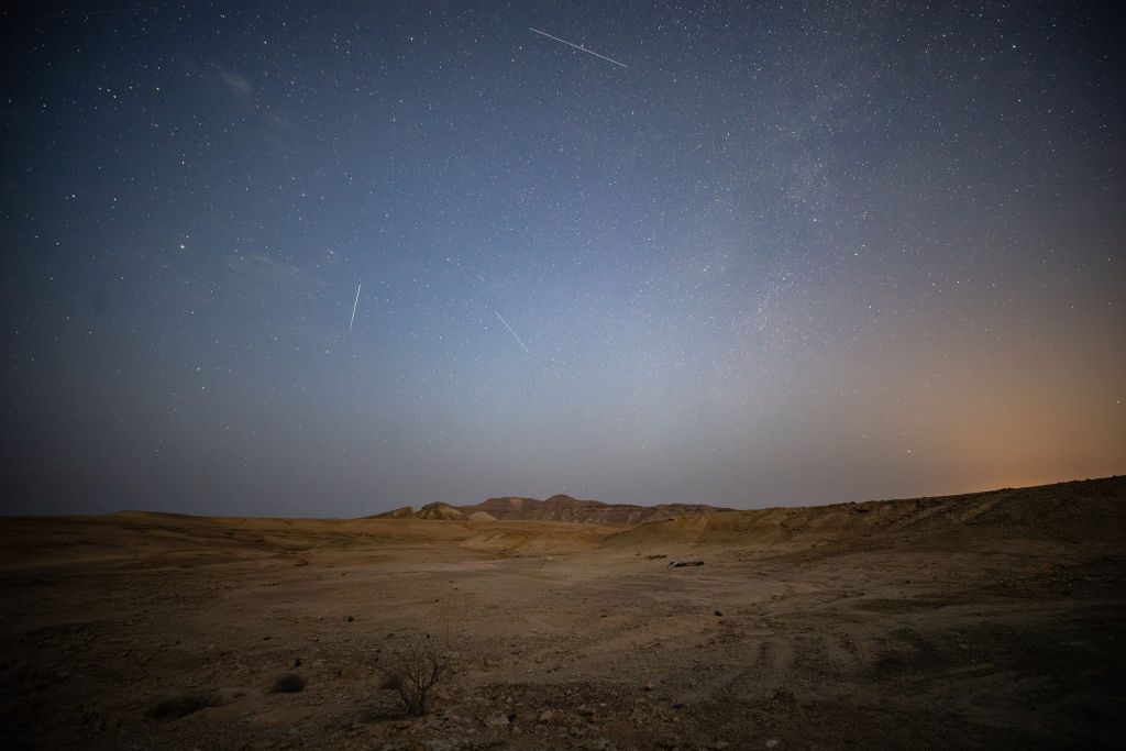 Banyak meteor melesat melintasi langit berbintang di atas lanskap gurun yang tandus.
