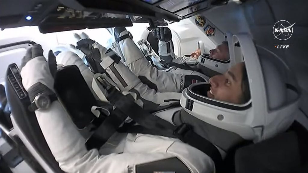 three astronauts in white spacesuits with open helmet faceplates sit inside a SpaceX capsule in orbit