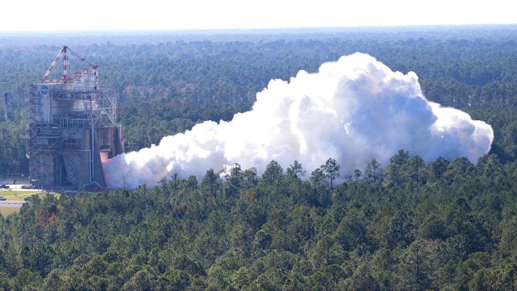 An industrial construct stands amidst a green forest with smoke billowing out into a plumeA full duration test of the RS-25 certification engine was conducted at NASA