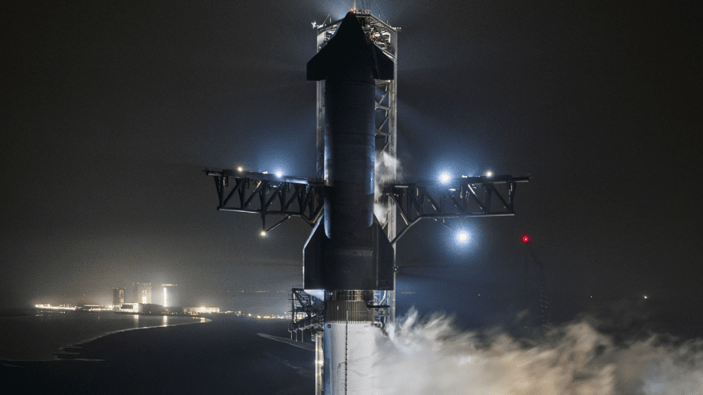 a large black and silver rocket vents vapor at night while standing upright on a launch pad