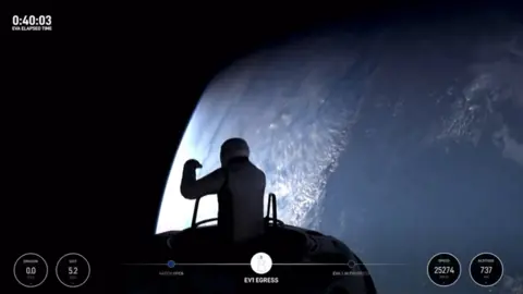 Jared Isaacman dari SpaceX muncul dari palka pesawat ruang angkasa pada awal perjalanan ruang angkasa pribadi pertama. Dia berada dalam bayang-bayang, mengenakan pakaian antariksa. Bumi dapat dilihat pada latar belakang di sisi kanan gambar. Itu adalah awan biru melengkung dan awan putih dapat dilihat.