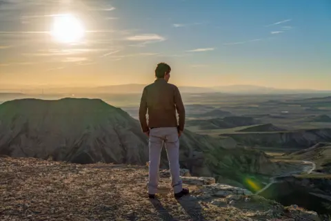 BBC Studios/Nick Gaven Profesor Brian Cox difilmkan dari belakang, memandang cakrawala dari puncak gunung. Dia mengenakan jaket coklat dan celana abu-abu. Perbukitan hijau terlihat di kejauhan dan matahari bersinar.