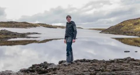 BBC Studios/Zach Levi-Rodgers Profesor Cox berdiri di atas bebatuan di tepi danau besar. Perbukitan terlihat di kejauhan. Dia mengenakan jaket abu-abu, celana dan sepatu berjalan, dan tersenyum.