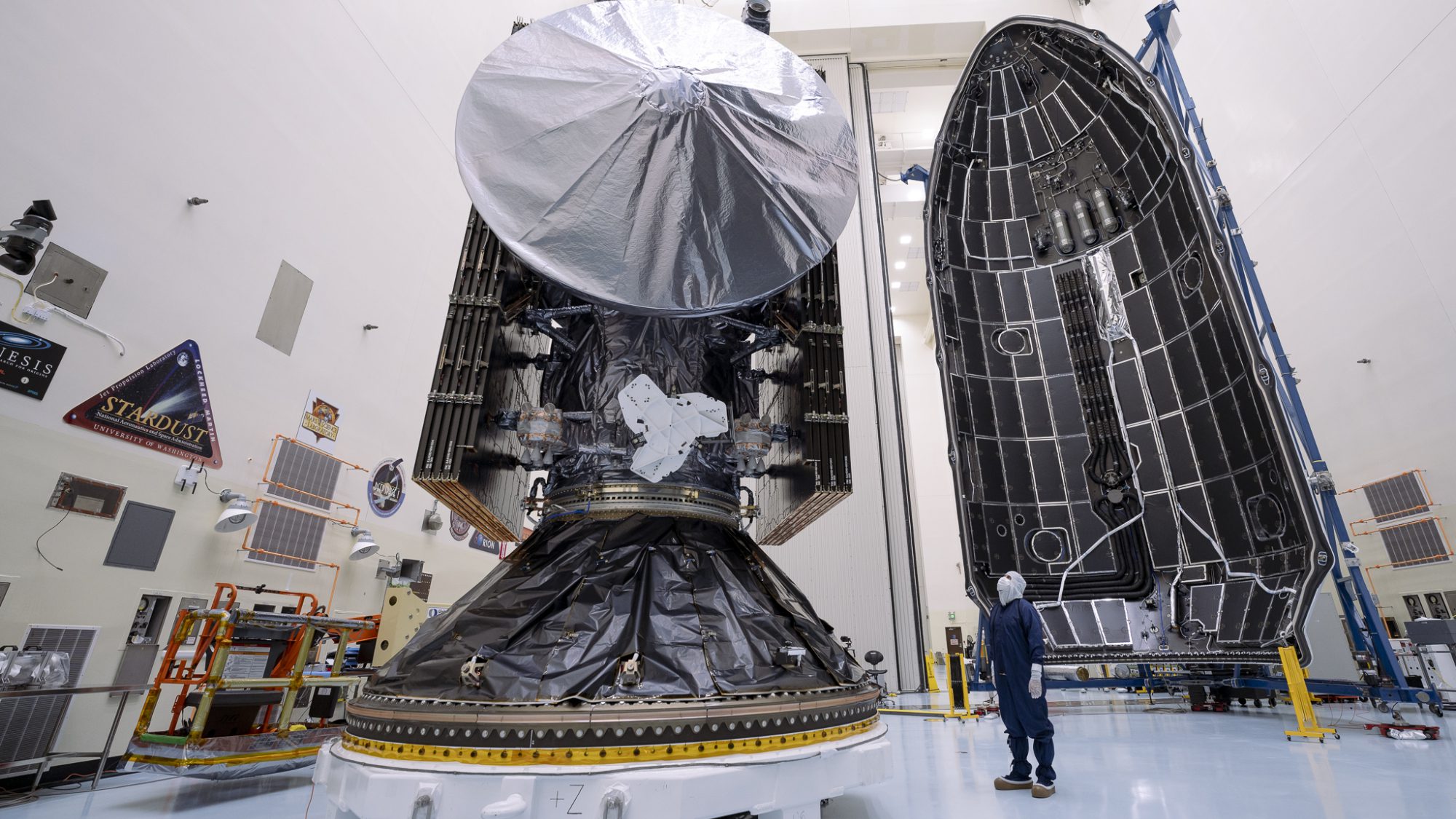 Pesawat luar angkasa Europa Clipper yang besar memiliki antena melingkar, seorang pria sedang melihatnya, dan kerucut hidung roket besar di latar belakang.