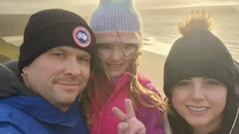 Angharad Denise Angharad Denise tersenyum saat berfoto selfie bersama suami dan putrinya di pantai. Mereka memakai topi dan mantel dengan matahari di belakang mereka.