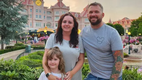 Angharad Dennis Angharad Dennis dan suaminya, Joe, mengenakan kaos Mickey Mouse berwarna abu-abu. Mereka berdiri di taman di Disneyland sambil tersenyum bersama putri mereka Carrie.