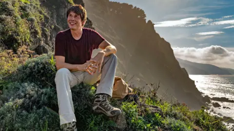 BBC Studios/Nick Gavin Profesor Brian Cox duduk di bukit yang tertutup pepohonan, dengan laut di sisi kanan gambar. Dia mengenakan kemeja merah anggur, celana abu-abu, dan sepatu berjalan. Dia tersenyum sambil melihat ke kejauhan. Matahari bersinar terang di latar belakang.